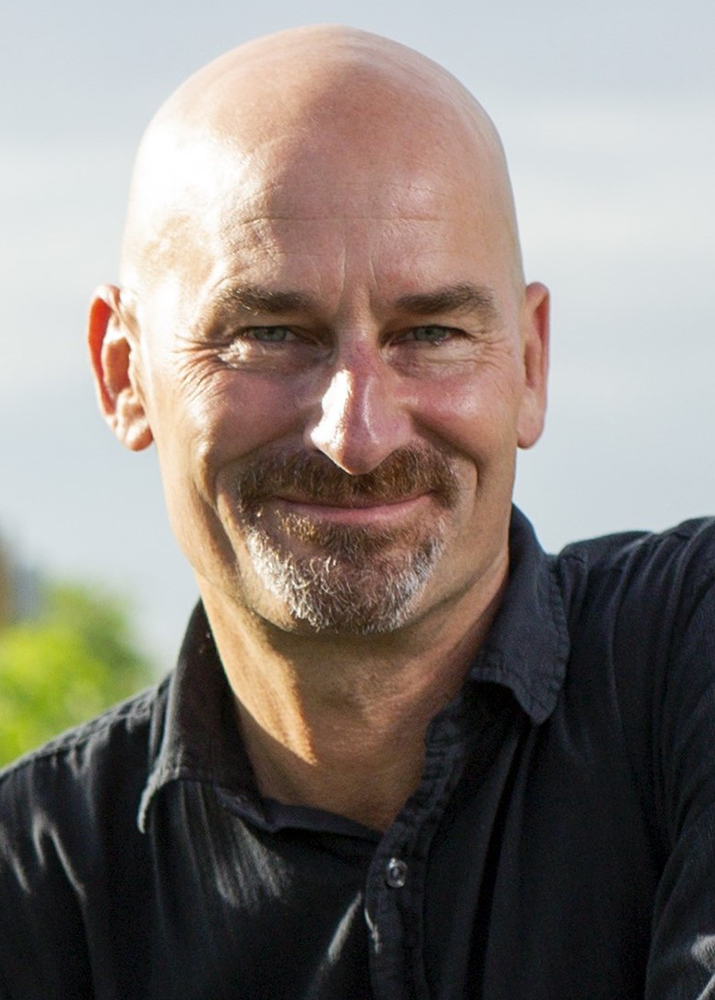 A man with a goatee is smiling outdoors, wearing a dark shirt. The background is blurred with greenery.